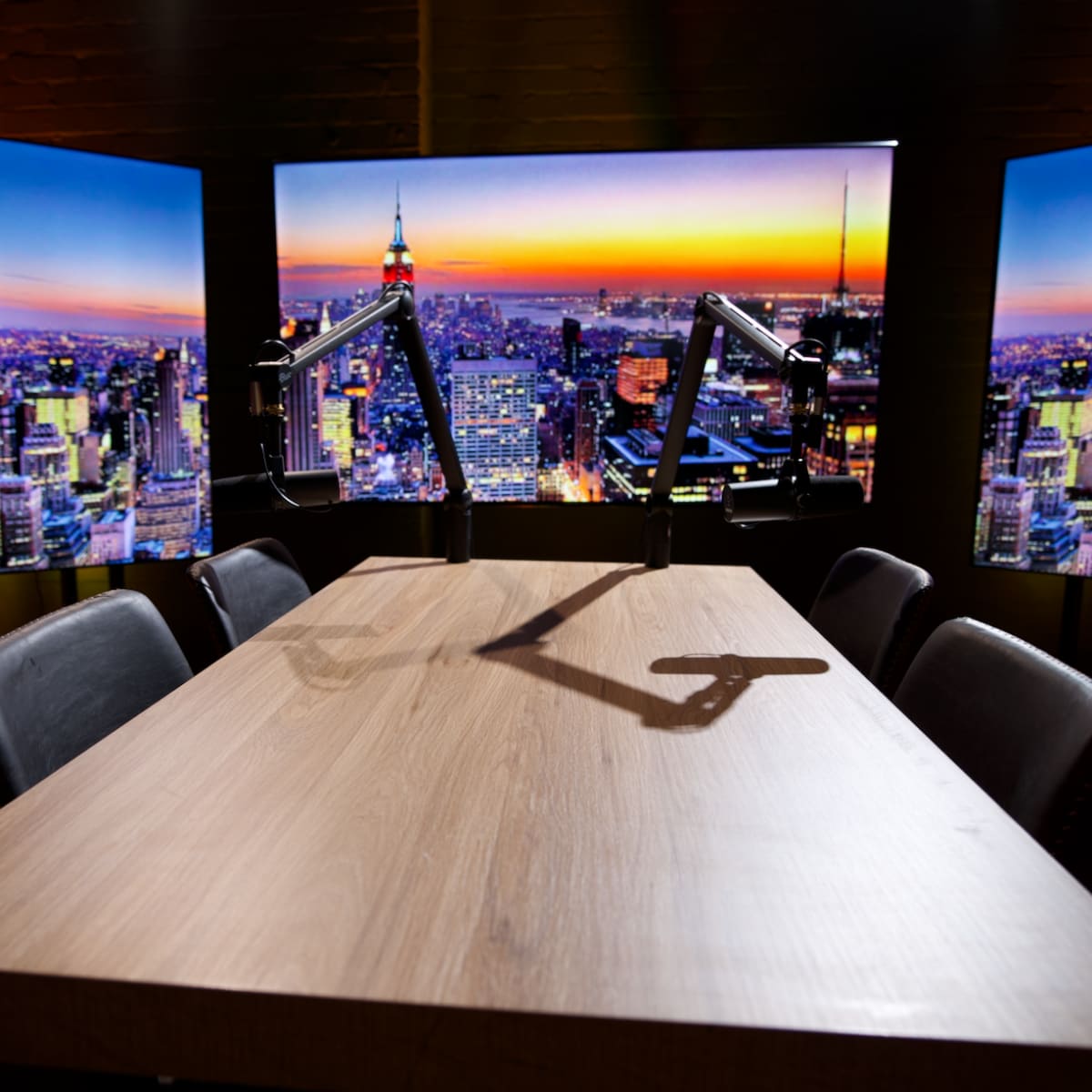 A modern conference room is set up with a large wooden table, equipped with microphone arms, and three screens displaying a city skyline at sunset.