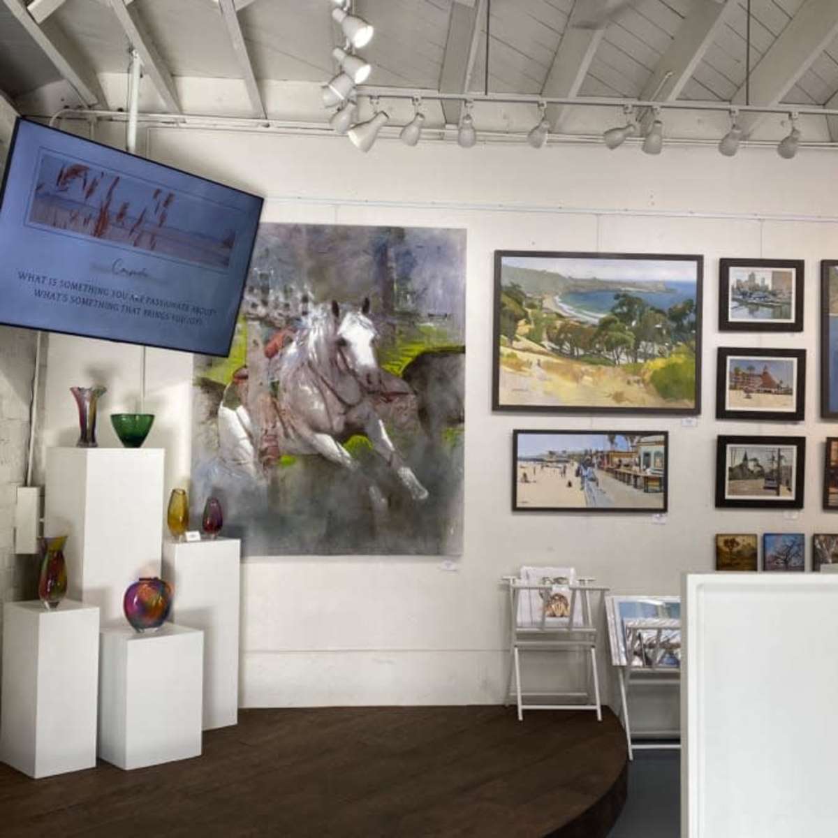 An art gallery interior with various paintings displayed on the walls and additional artworks arranged on shelves.