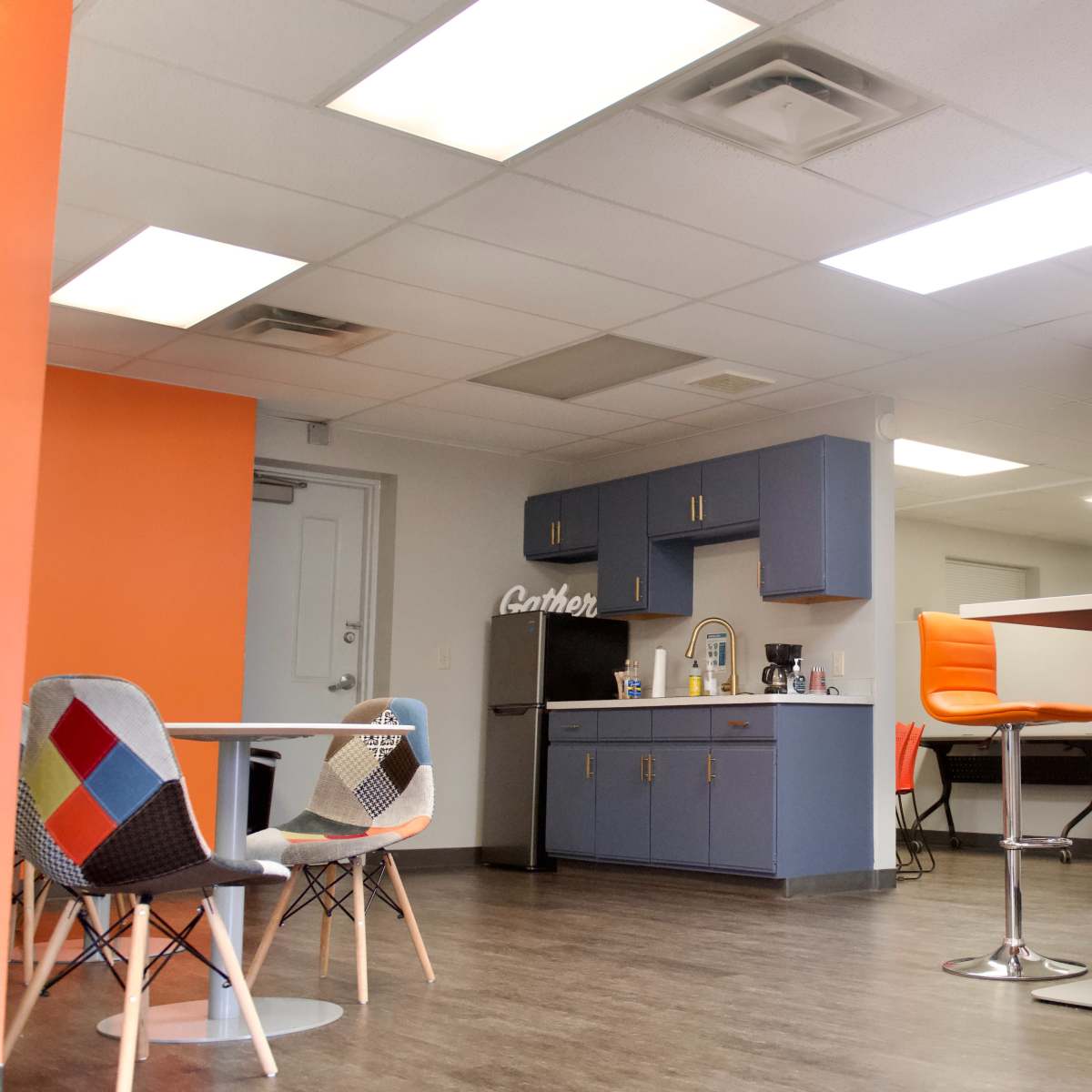 A modern office kitchen area with blue cabinets, a black refrigerator, orange accent walls, and various seating options including colorful chairs and bar stools.
