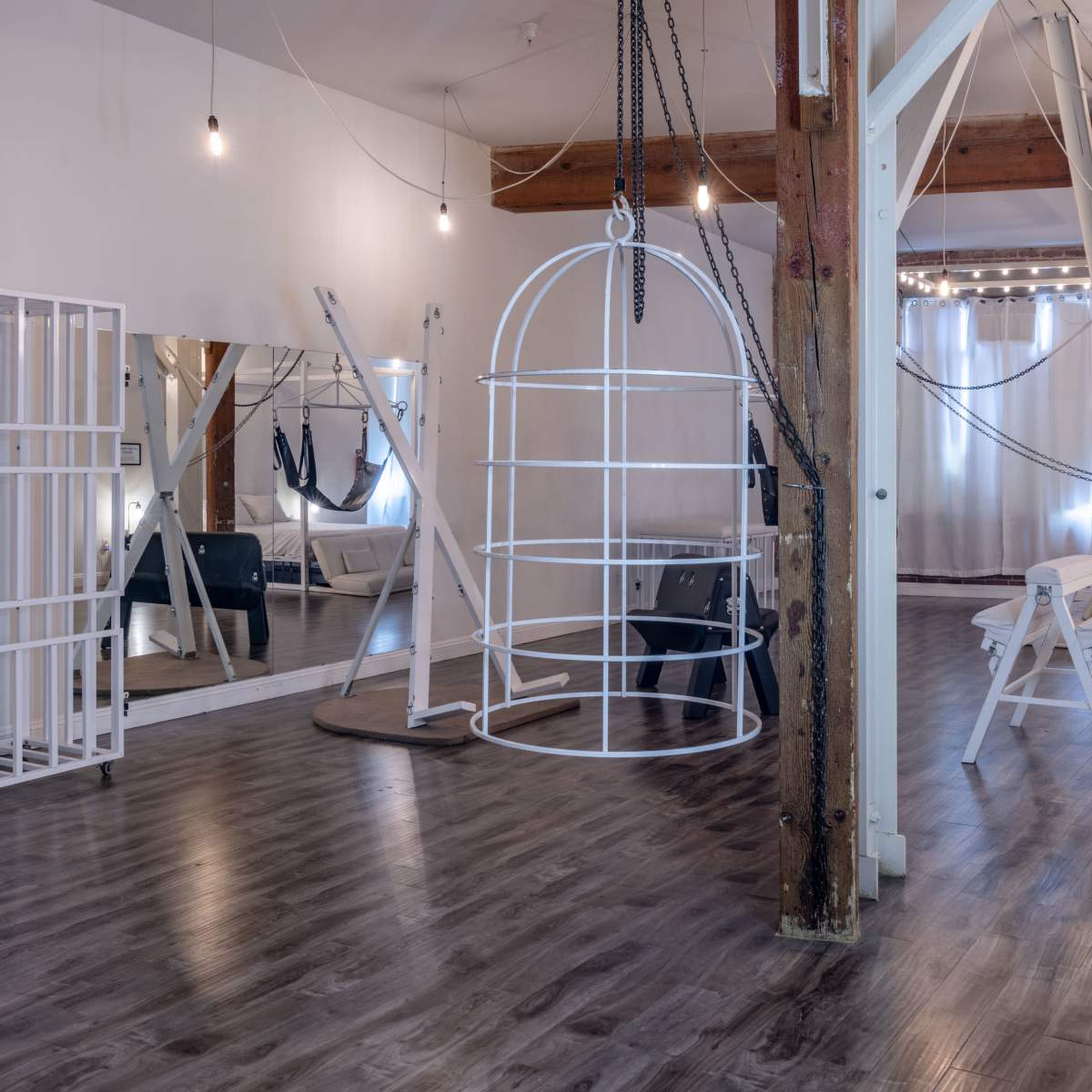 A spacious, minimalist studio with wooden beams, white furnishings, a hanging structure, and a large mirror.