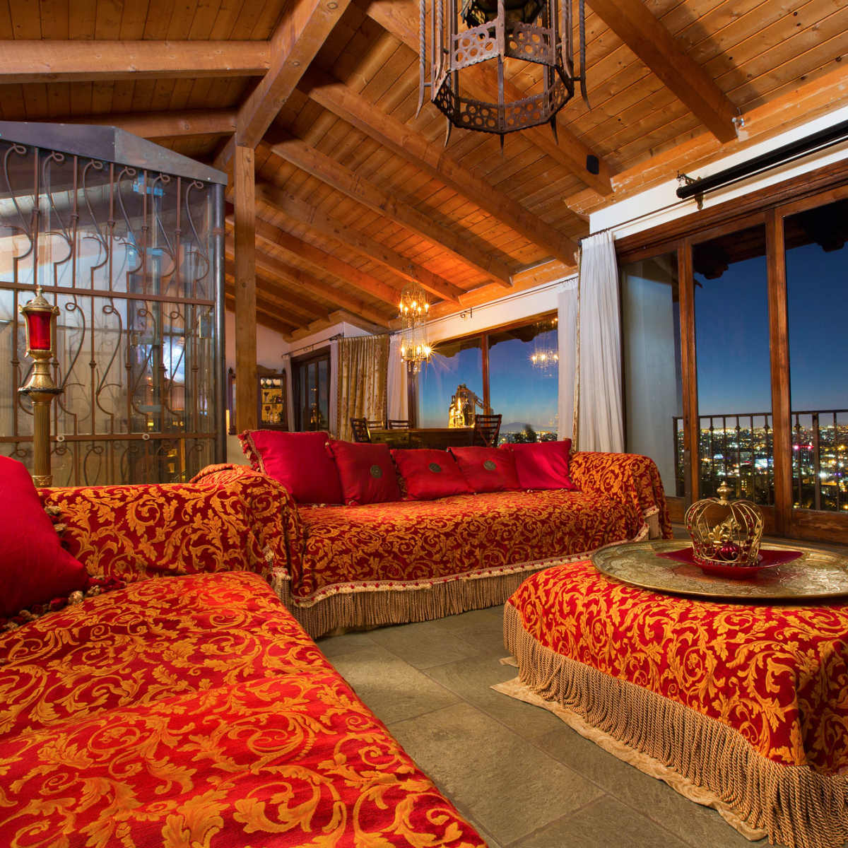 A spacious living room with red and gold patterned sofas, a decorative coffee table, and large windows showcasing a city skyline at dusk.