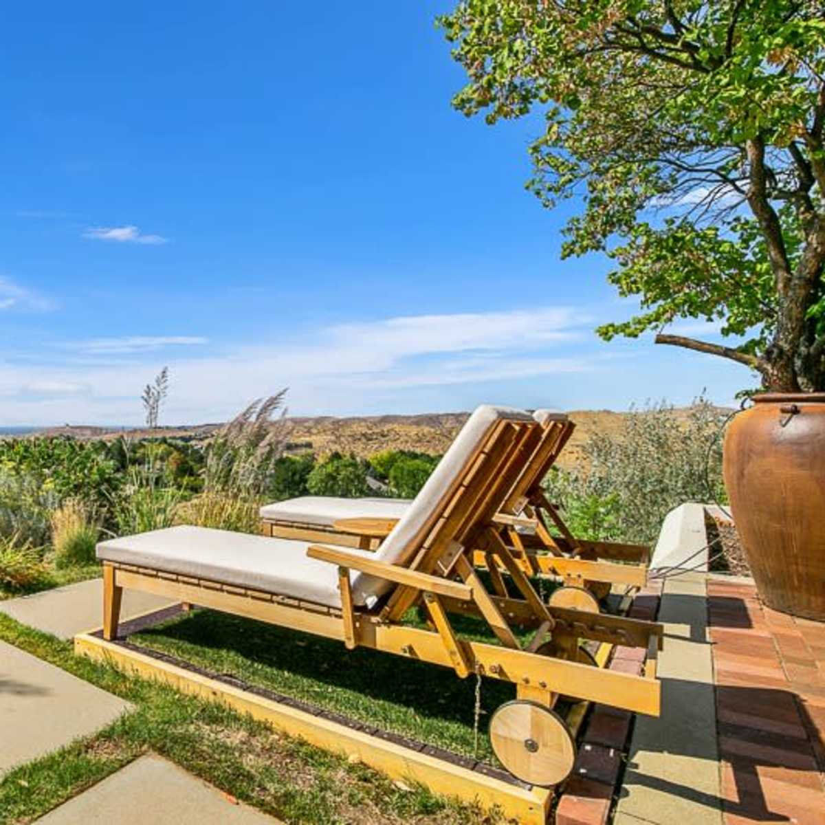 Two lounge chairs are positioned on a patio overlooking a vast landscape of hills and greenery under a clear blue sky.