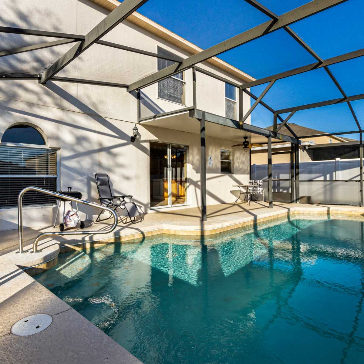 A swimming pool surrounded by a screened enclosure in a residential backyard, with a lounge chair and small table visible nearby.