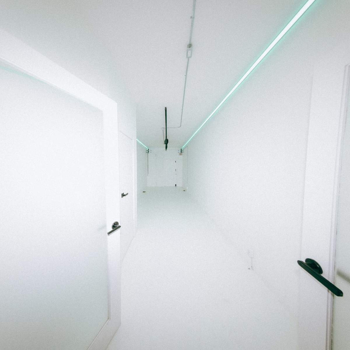 A long, narrow corridor with white walls and floors, featuring several closed doors and illuminated by a strip of blue-green light along the ceiling.