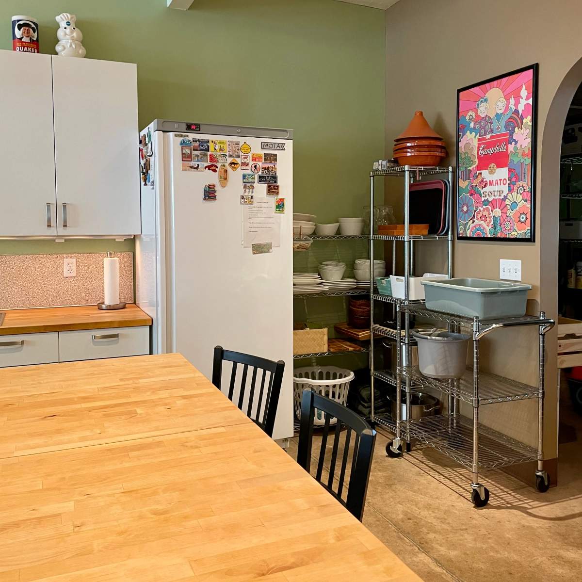 A kitchen with a wooden dining table, a refrigerator covered in magnets, and shelves with various kitchenware against a green wall.