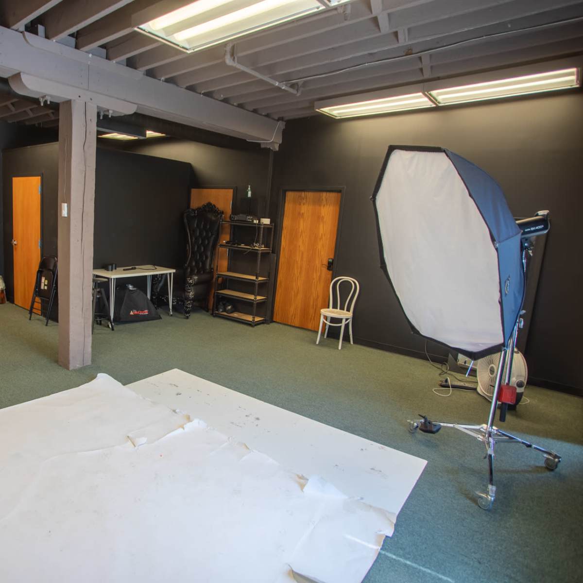 A photography studio with a large softbox light, a white backdrop on the floor, and several wooden doors against dark walls.