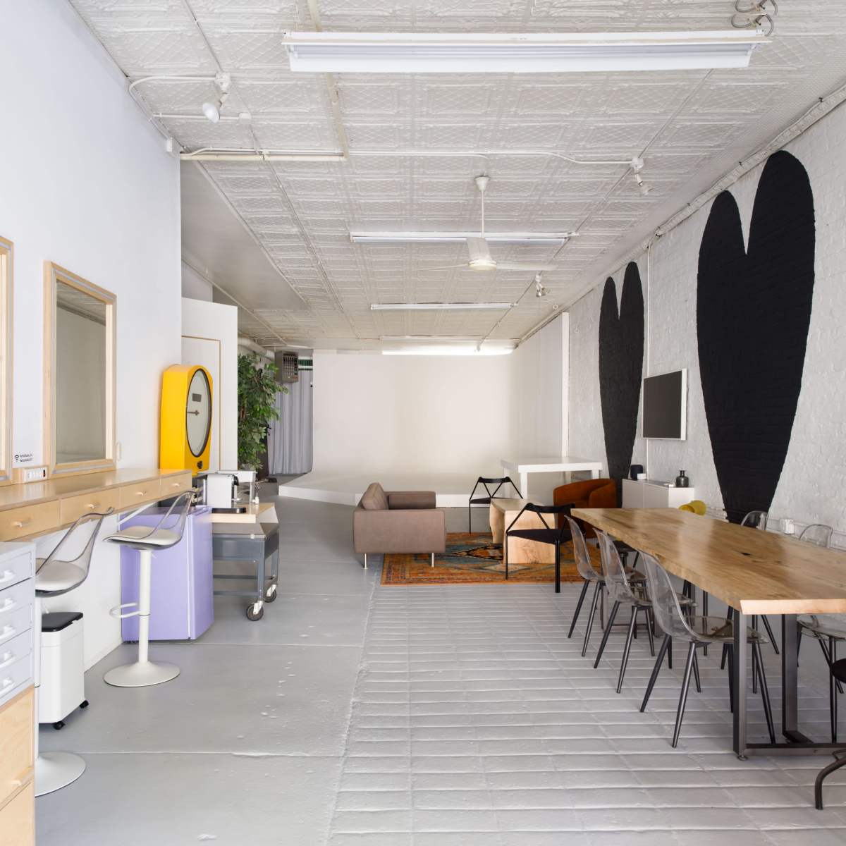 A spacious interior features large black heart murals on the walls, a wooden dining table, a sofa, and several workstations.