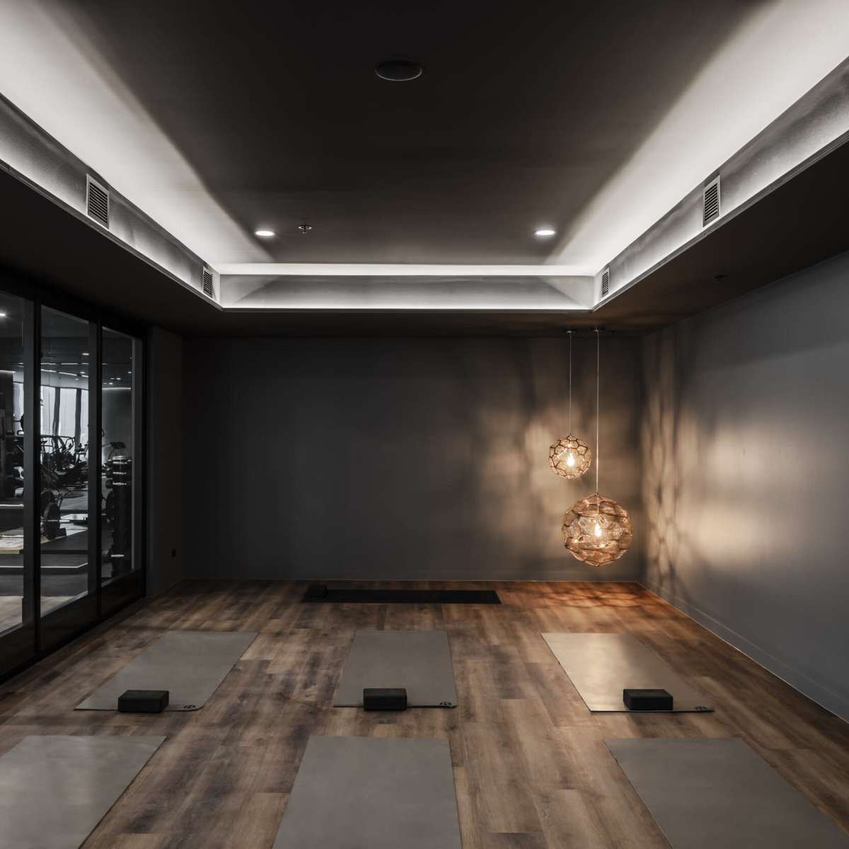 A dimly lit yoga studio features two hanging light fixtures and several yoga mats aligned on a wooden floor, with large windows revealing a gym area in the background.