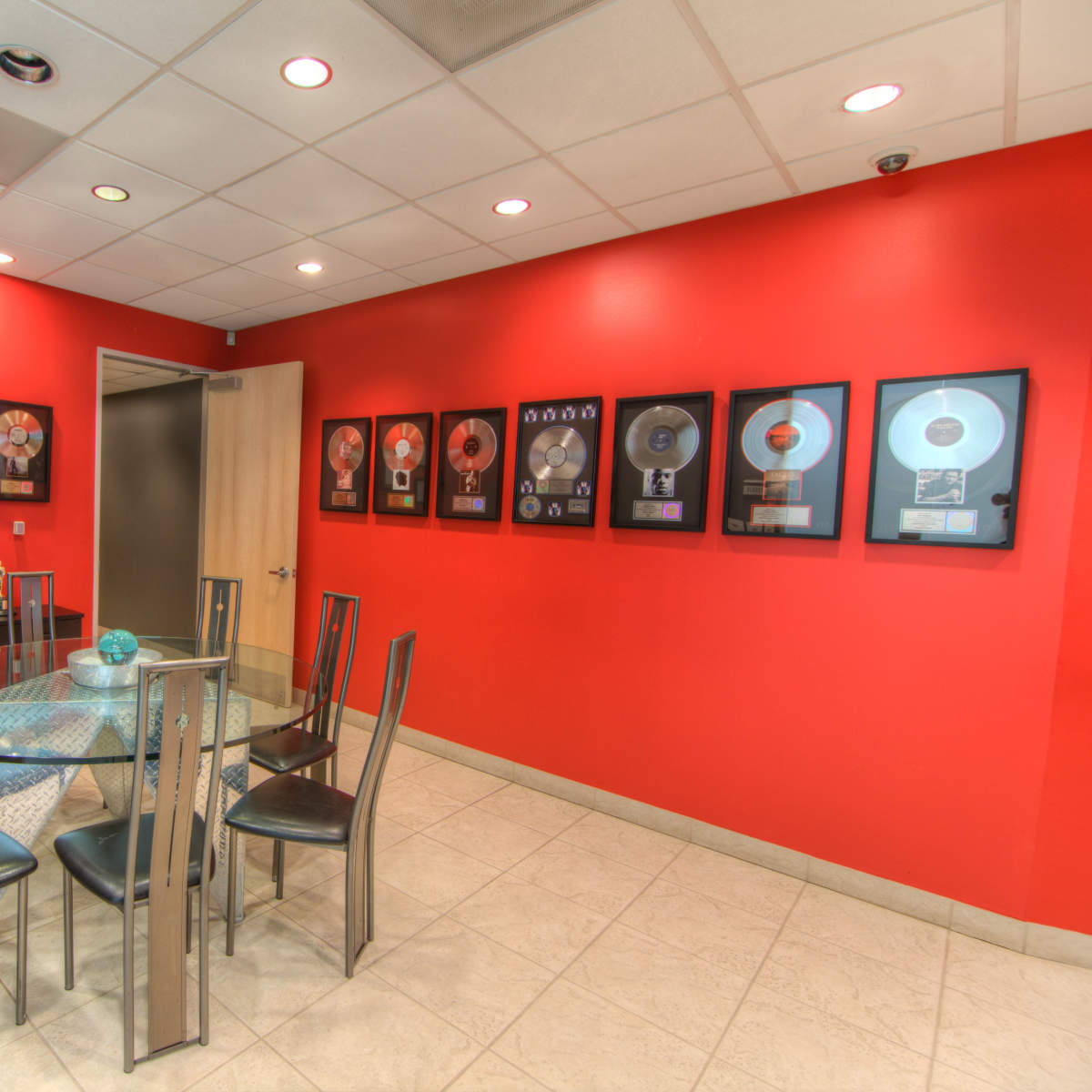 A conference room featuring a glass table surrounded by black chairs, with red walls displaying framed music awards and certificates.