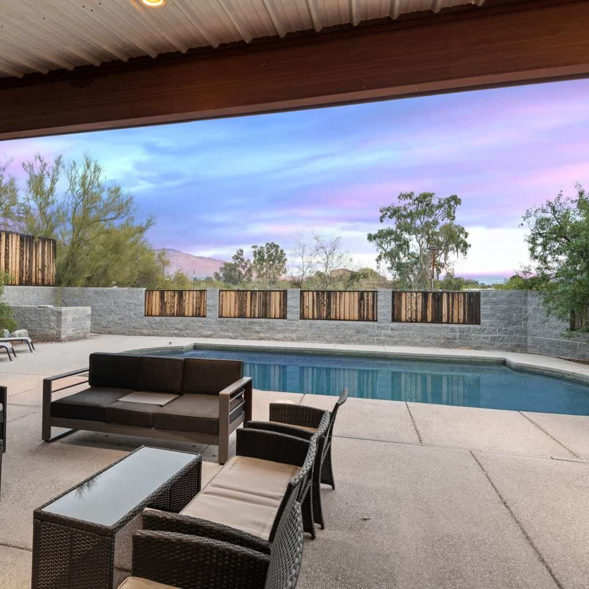 A backyard with a swimming pool, outdoor seating, and a fence surrounded by greenery under a colorful sunset sky.