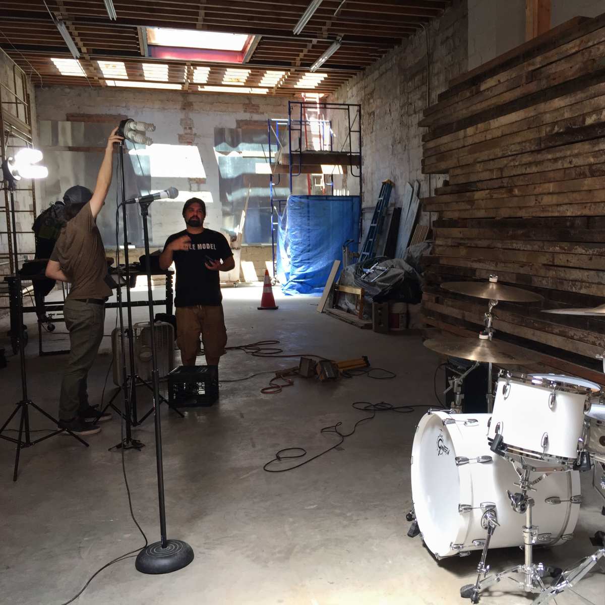 A musician stands with a microphone in a spacious, unfinished warehouse, next to a drum set, while a crew member adjusts lighting equipment.