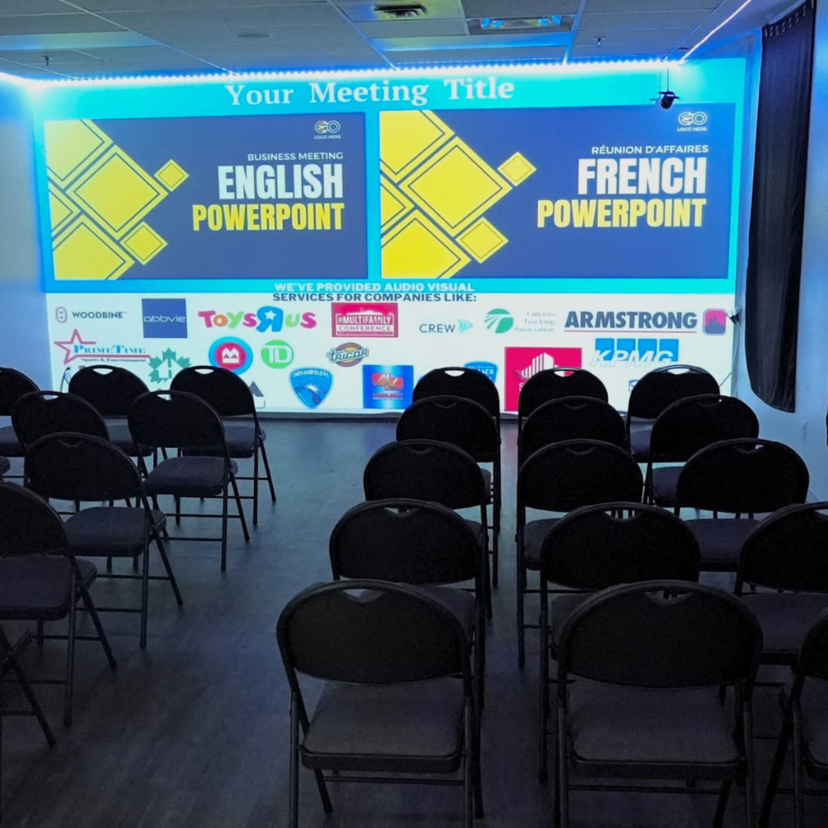 A meeting room is set up with rows of chairs facing two large screens displaying "English PowerPoint" and "French PowerPoint," with various sponsor logos on the walls.