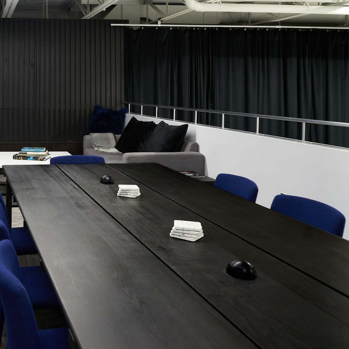 A long black conference table with blue chairs is positioned in a modern meeting room featuring a gray sofa and a dark wall with curtains.