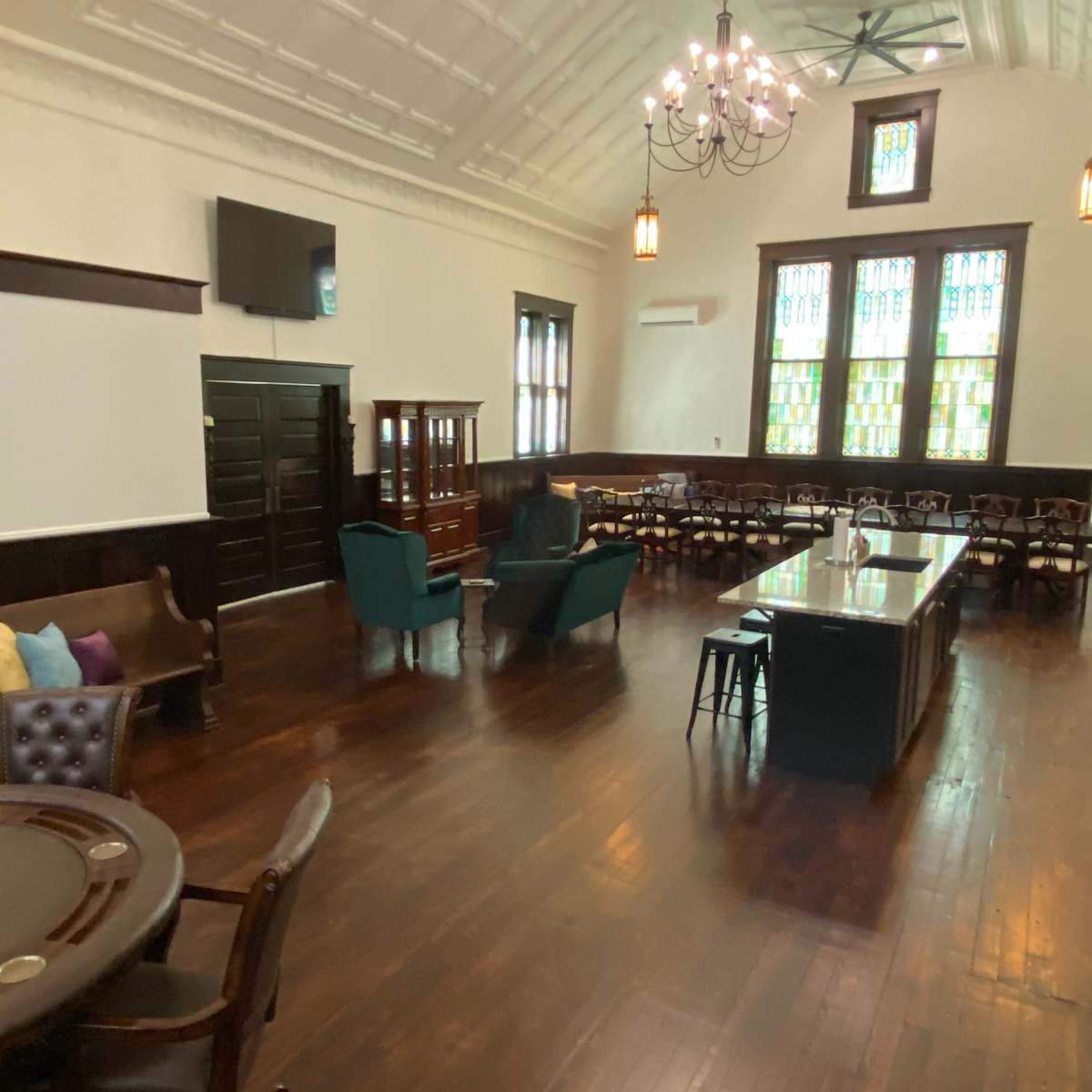 A spacious interior room with wooden floors, a large table, several seating areas, and stained glass windows.