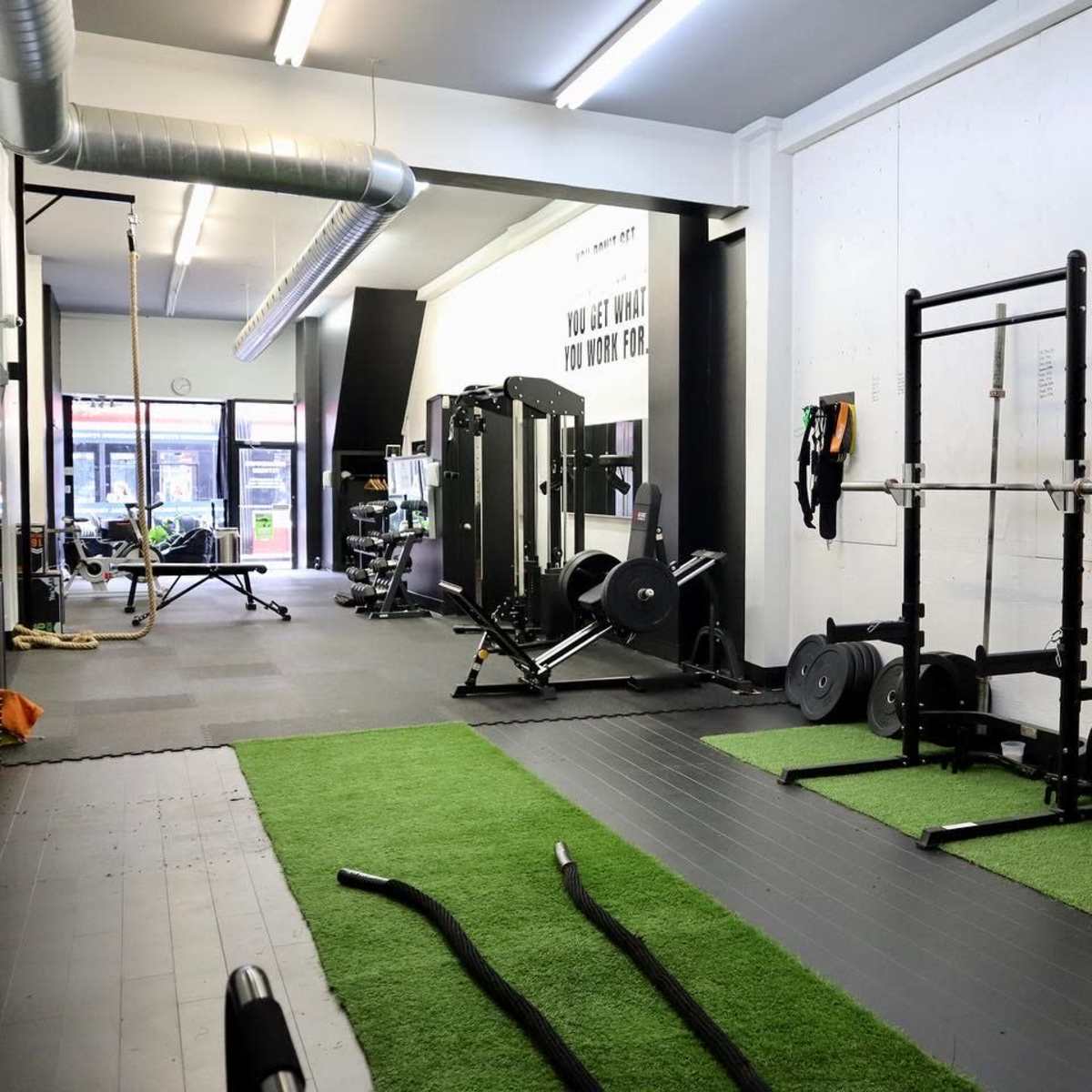 A gym interior with various exercise equipment, including weightlifting racks, a rowing machine, and training mats on a green carpet.