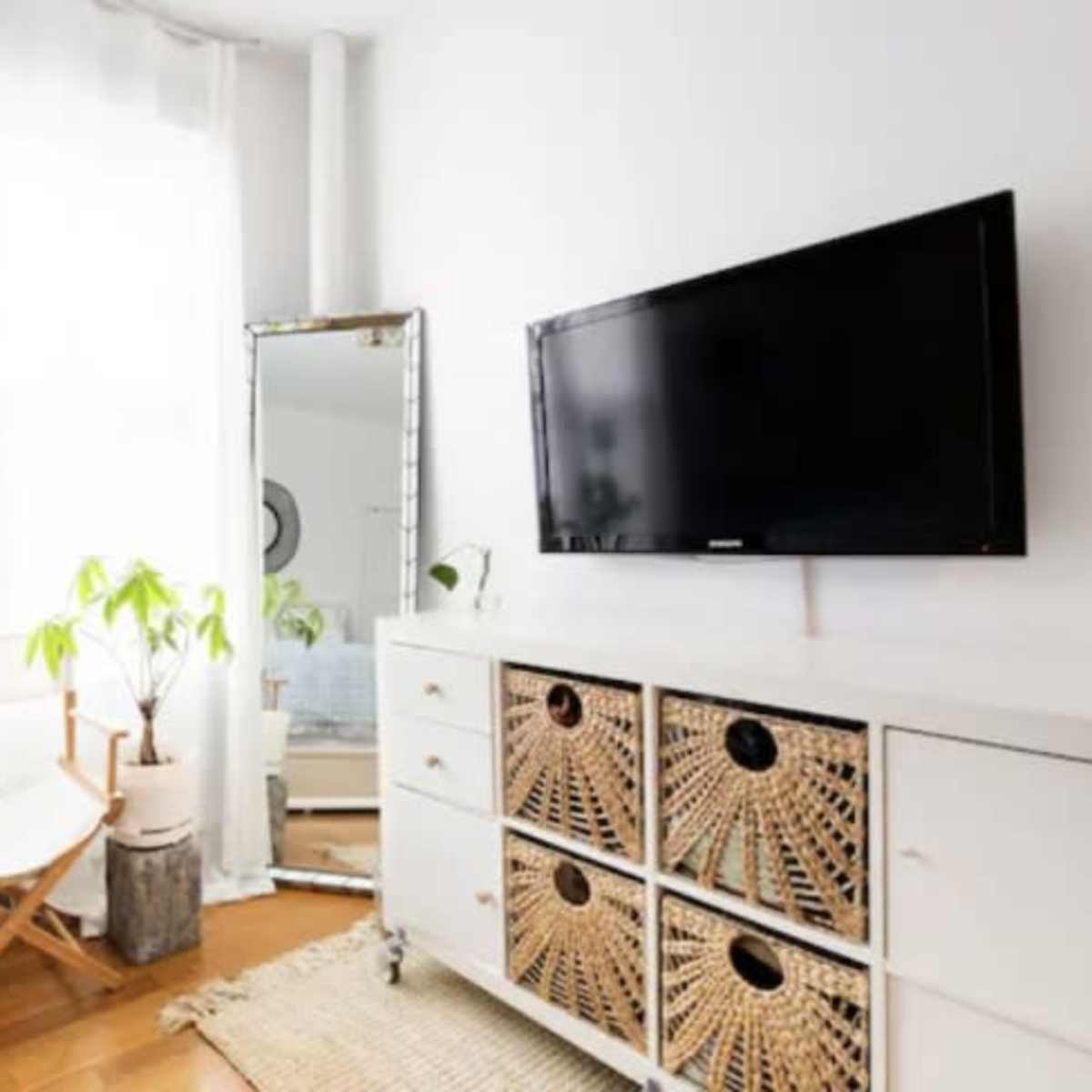 A bright, modern living space featuring a television mounted on a wall above a white cabinet with woven baskets, alongside a large mirror and potted plants.