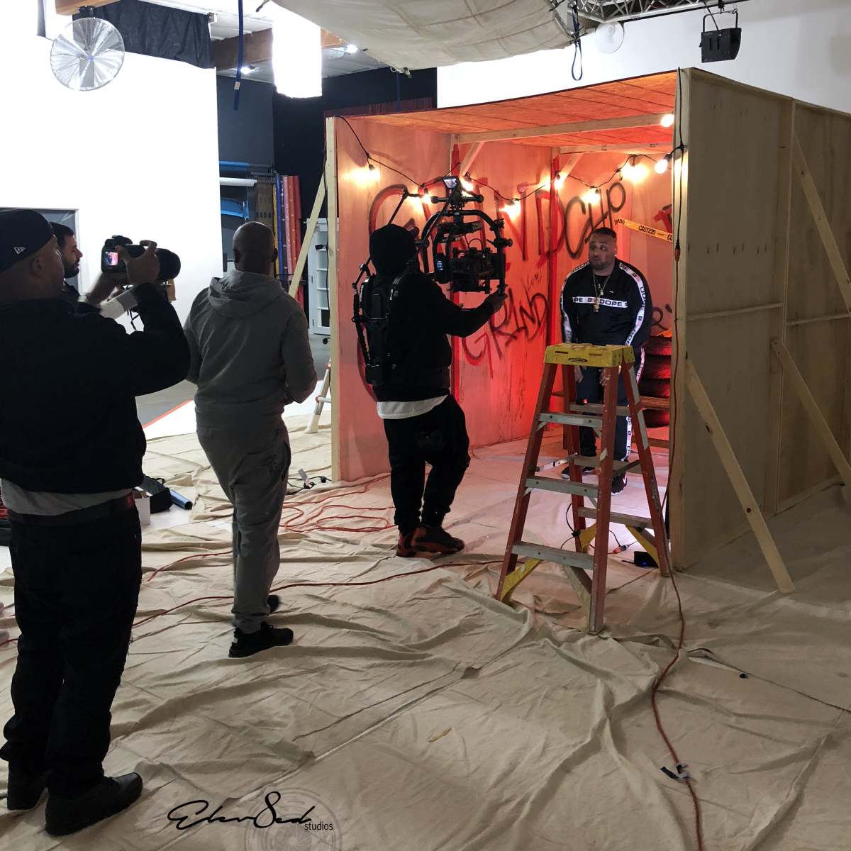 A film crew is setting up a scene inside a wooden structure, with a ladder in the foreground and lights illuminating the set.