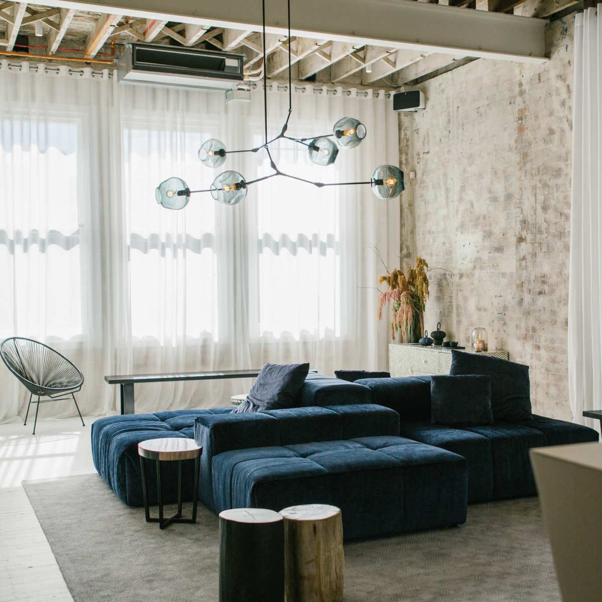 A modern living room with a large blue sectional sofa, a glass chandelier, and tall windows draped with sheer curtains.