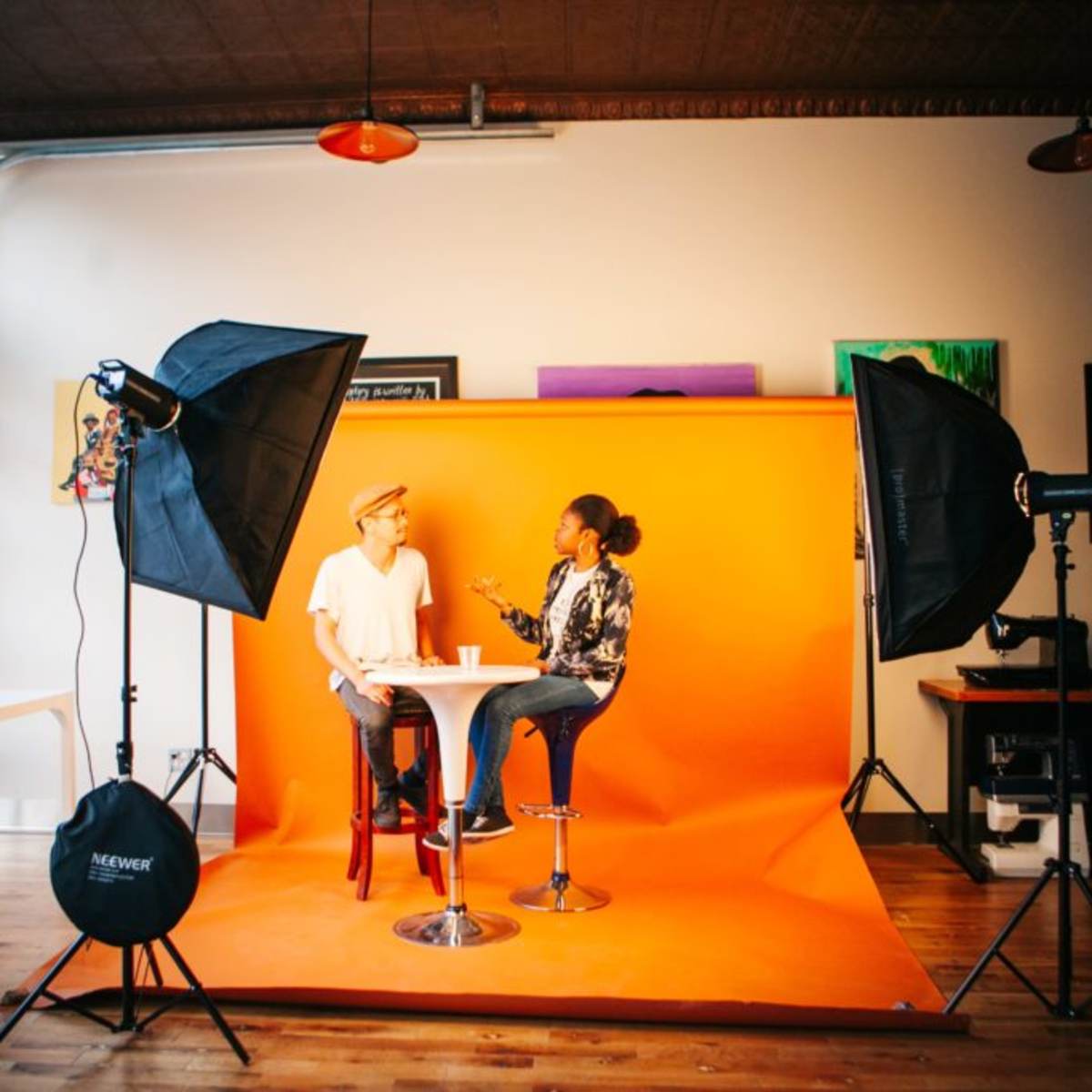 Two individuals are seated at a round table in front of an orange backdrop, surrounded by studio lighting equipment.