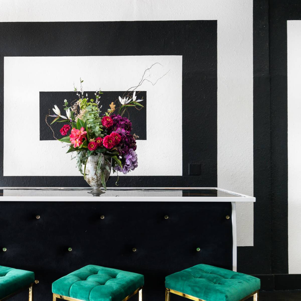 A black and white interior features a sleek table adorned with a large floral arrangement, flanked by green stools and a mannequin.
