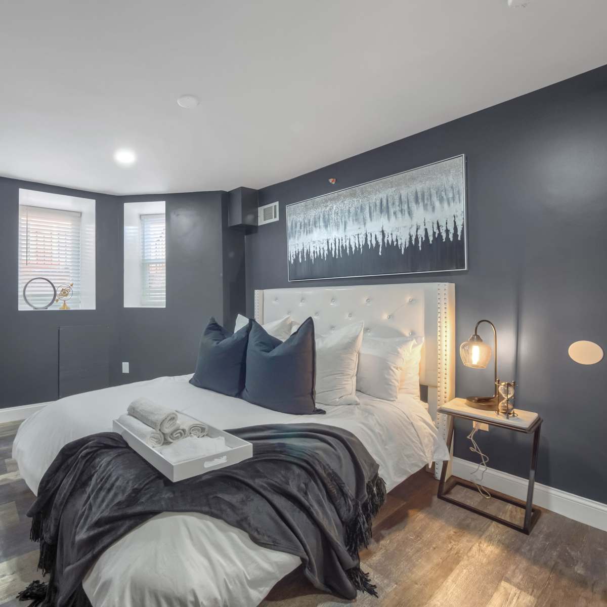 A modern bedroom with a bed dressed in white linens, dark walls, and a small side table with a lamp.