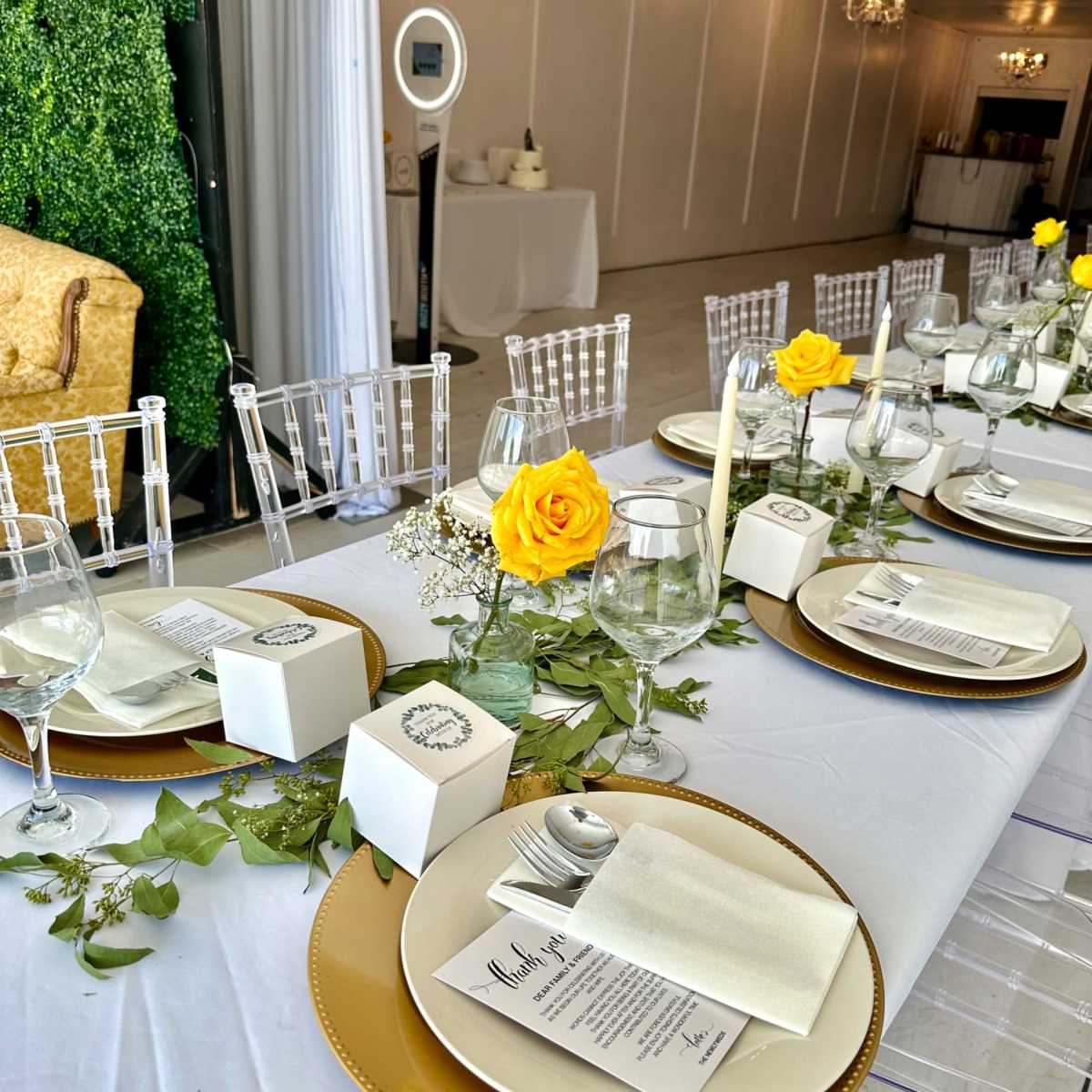 A elegantly set dining table with white tablecloths, gold chargers, glassware, and yellow roses as centerpieces.