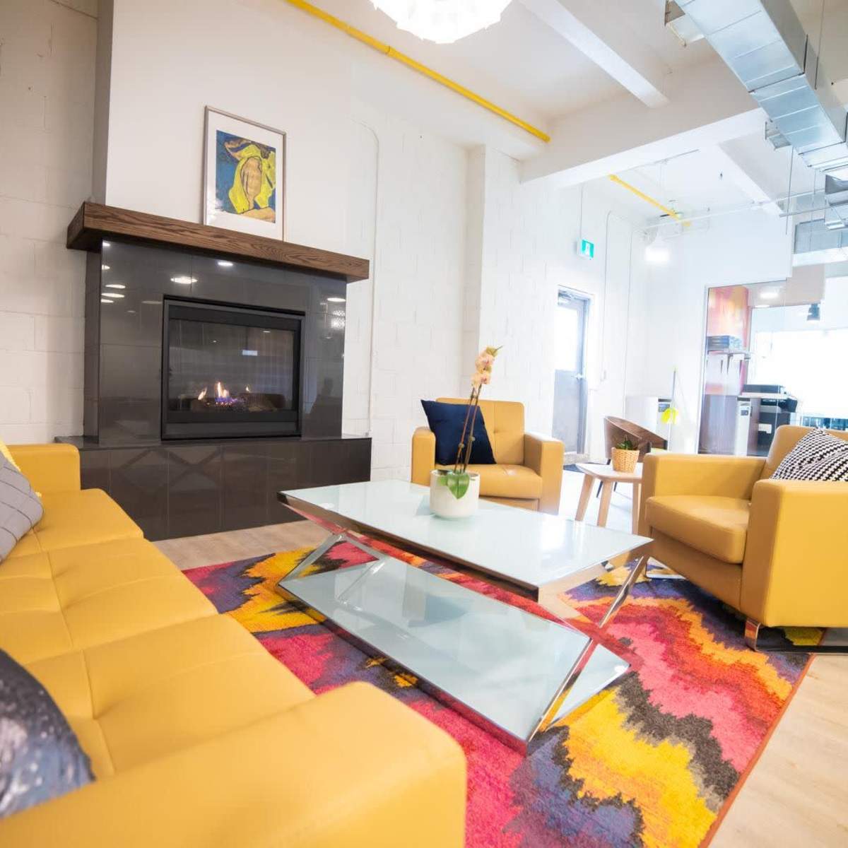 A modern living room with a yellow leather sofa, two yellow armchairs, a glass coffee table, a colorful rug, and a wall-mounted fireplace.