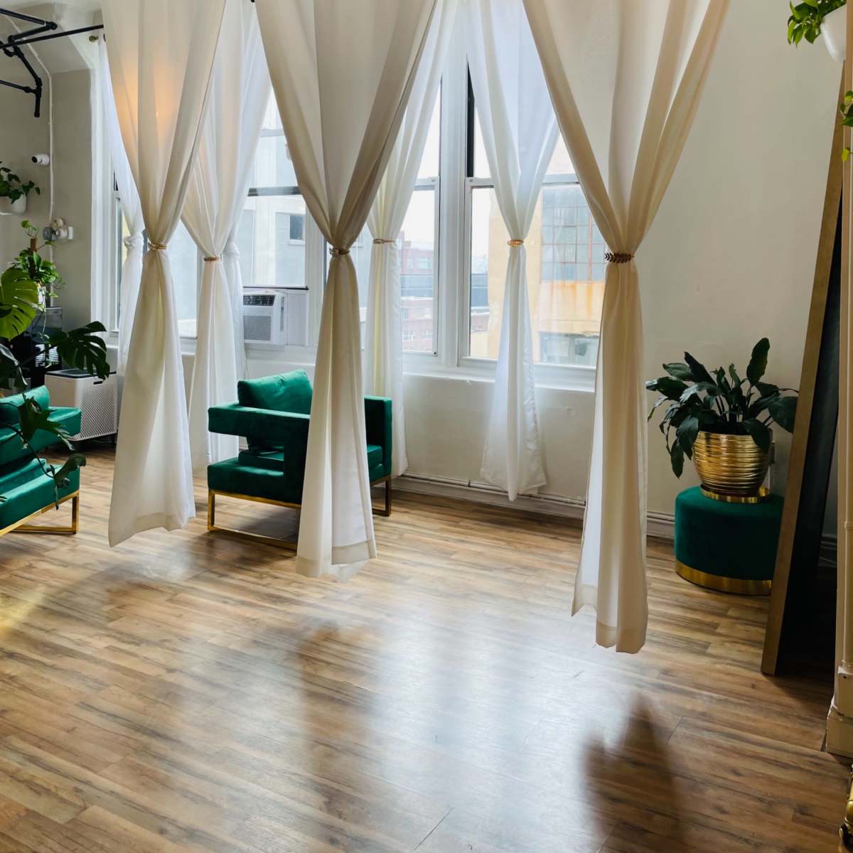 A bright room with large windows, decorated with white curtains and green upholstered chairs, along with potted plants.
