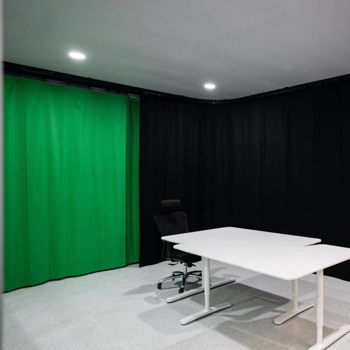 A small studio space features a green screen, black curtains, a white desk, and a black chair.