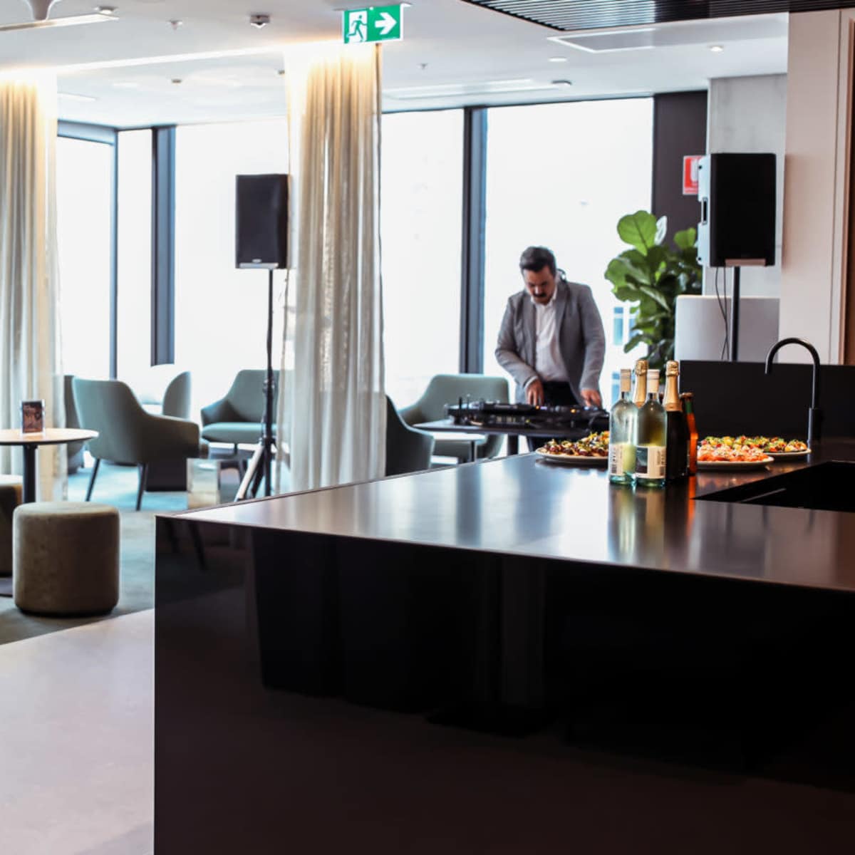 A modern lounge area features a curved green seating arrangement, tables, and a man playing music while others converse in the background.