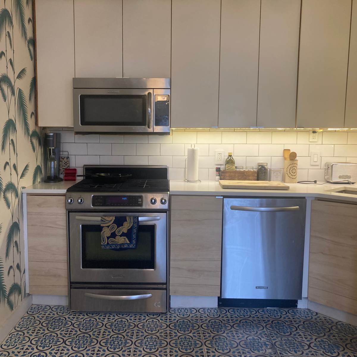 The kitchen features stainless steel appliances, including a stove and microwave, alongside light wood cabinetry and a decorative blue and white tile floor.