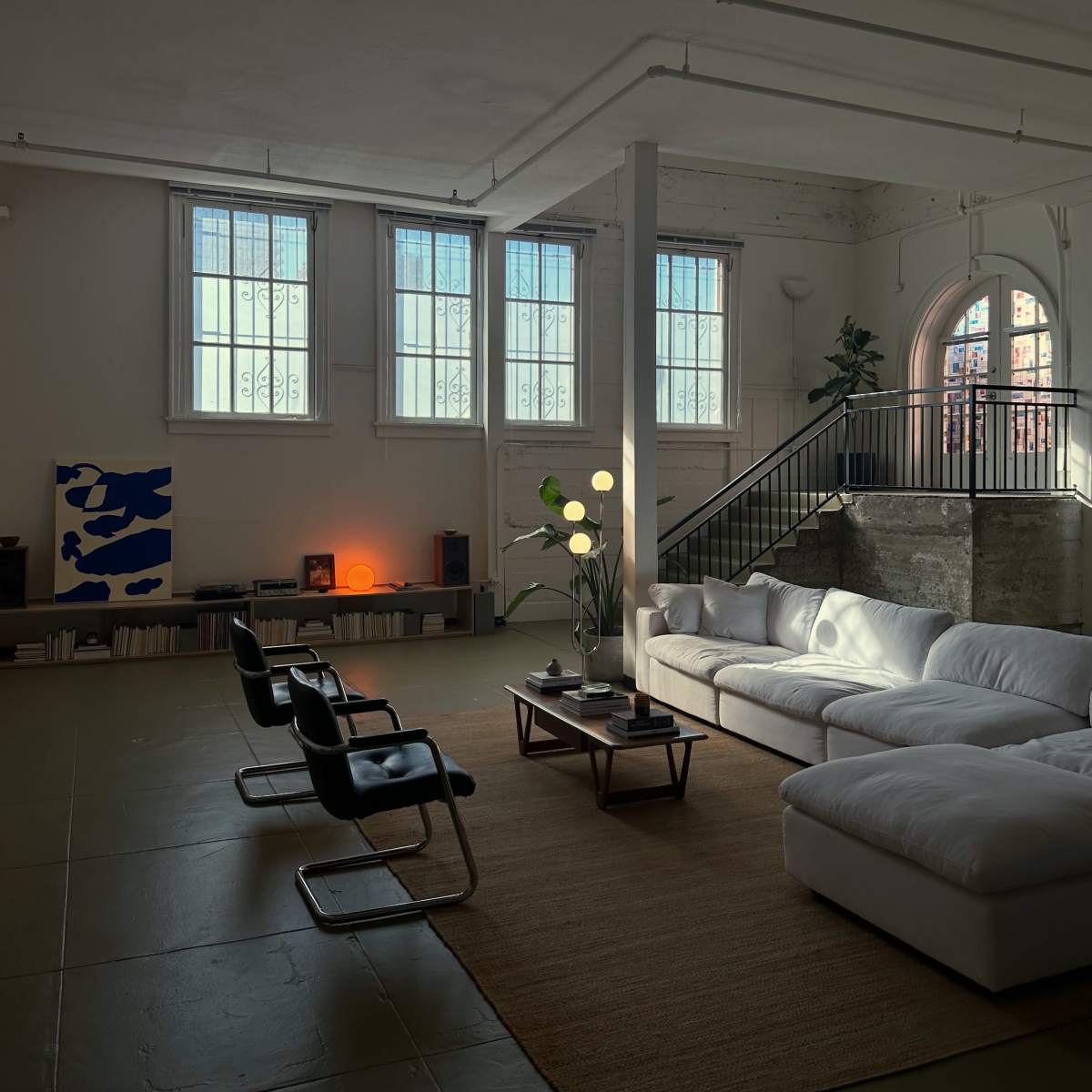 A cozy living area with a large white sectional sofa, two black chairs, a coffee table, and floor lamps, set against a backdrop of tall windows and concrete walls.