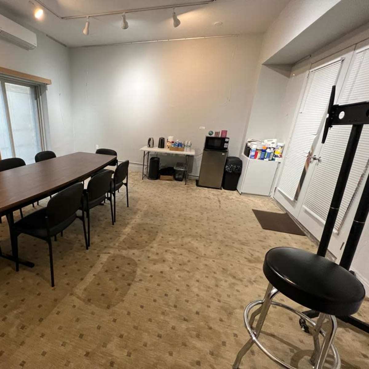 A conference room features a long table surrounded by chairs, with a kitchenette area and a large window in the background.