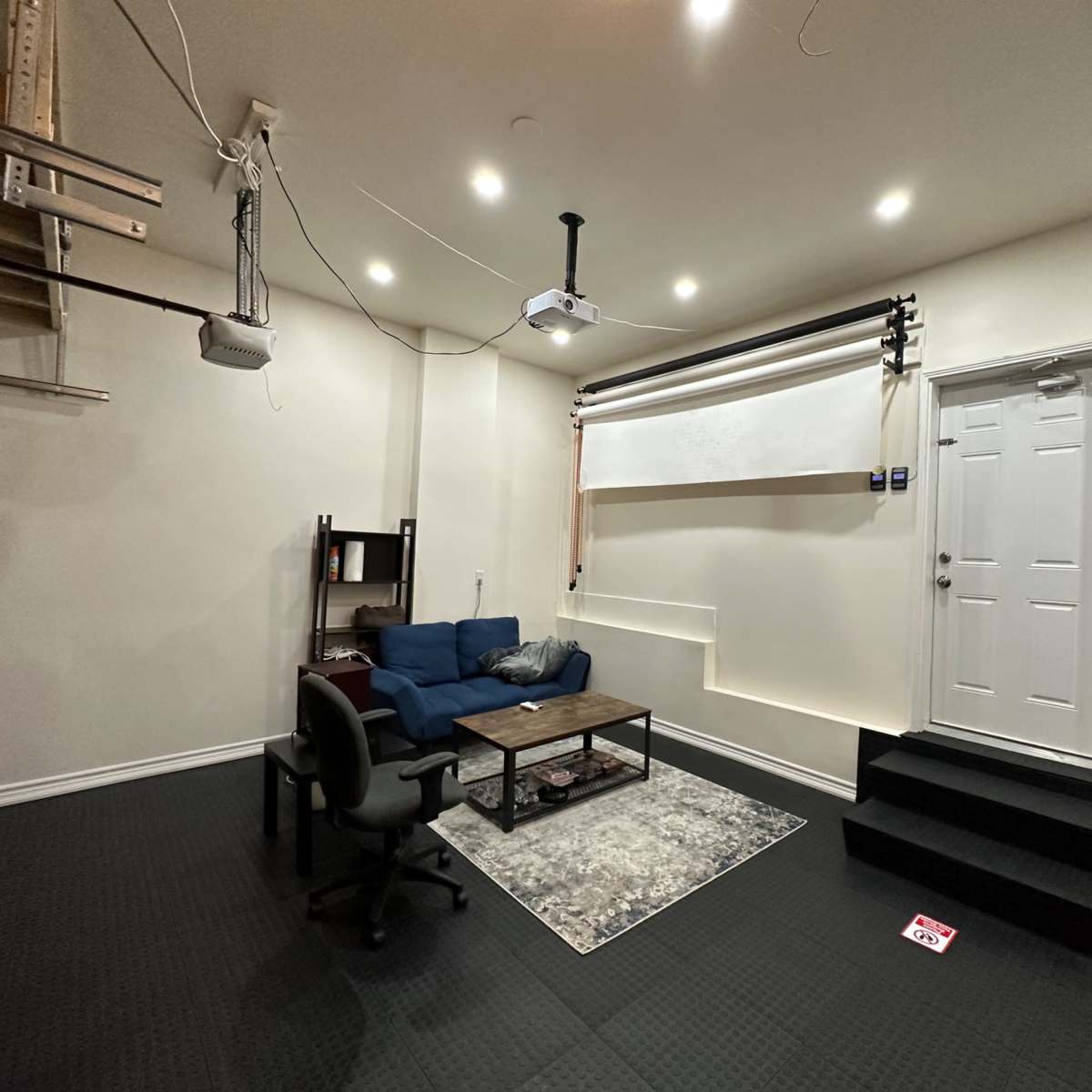 A sparsely furnished room with a blue couch, a coffee table, and a door leading outside, illuminated by overhead lights.