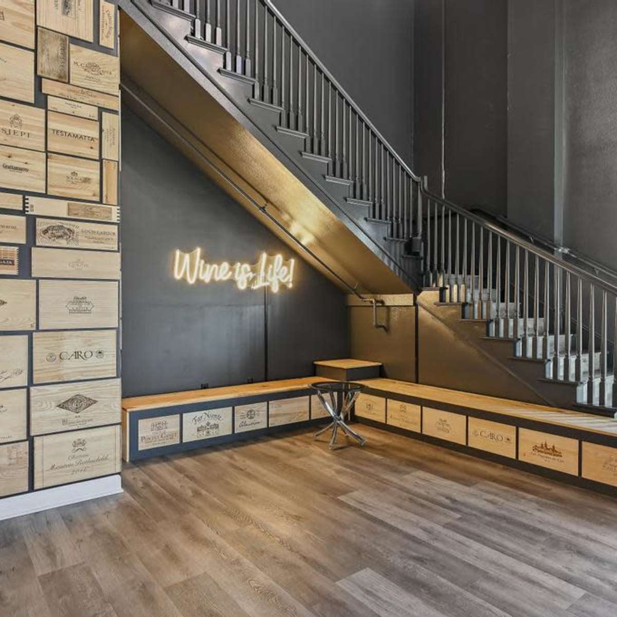 A modern interior space featuring a wooden wall adorned with wine crate labels and a staircase leading to an upper level, with a neon sign that reads "Wine is Life."