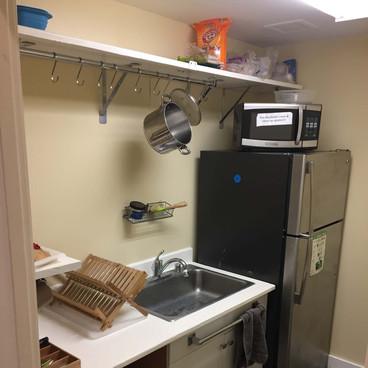 A compact kitchen area featuring a sink, a black refrigerator, a microwave on a shelf, and various kitchen utensils hanging above.