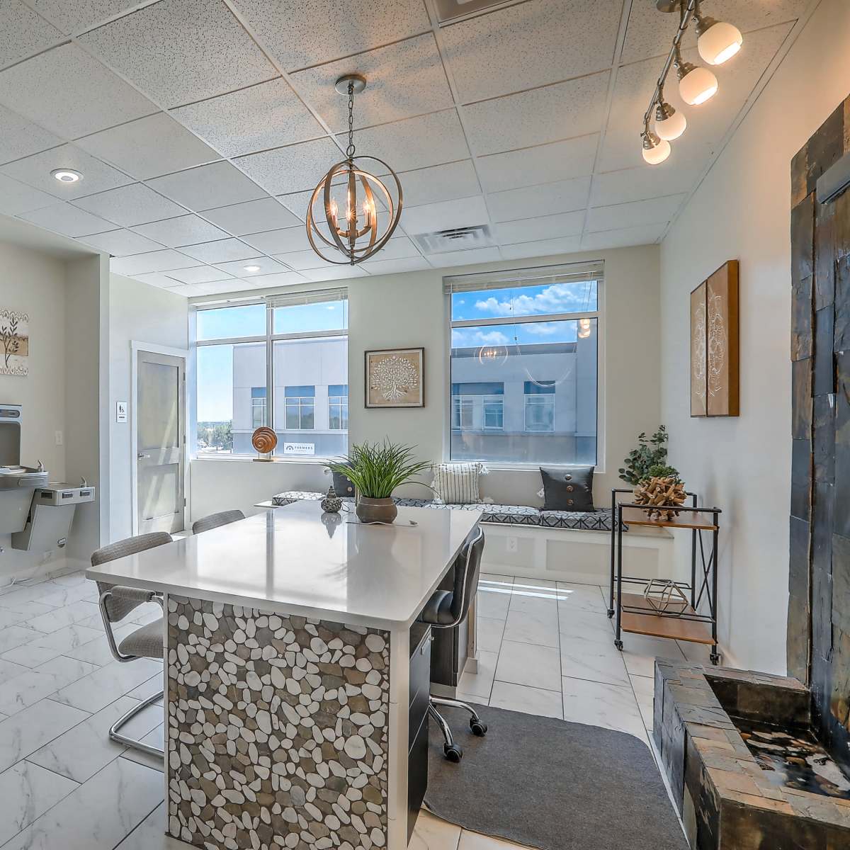 A modern office space featuring a large island desk with a mosaic design, windows allowing natural light, and a minimalist decor.