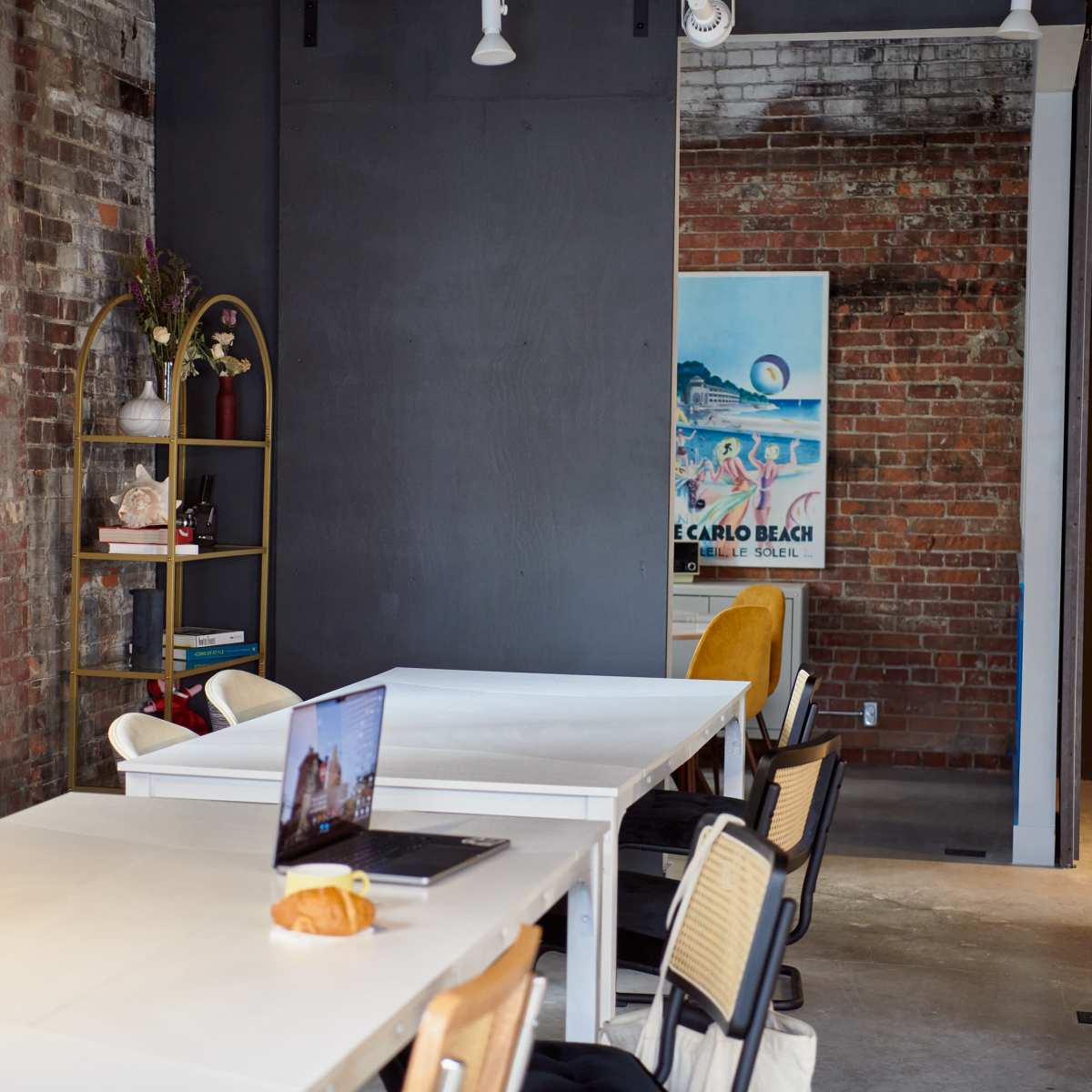 A modern workspace with a white table, chairs, and a bookshelf against a brick wall, featuring a laptop and a croissant on the table.