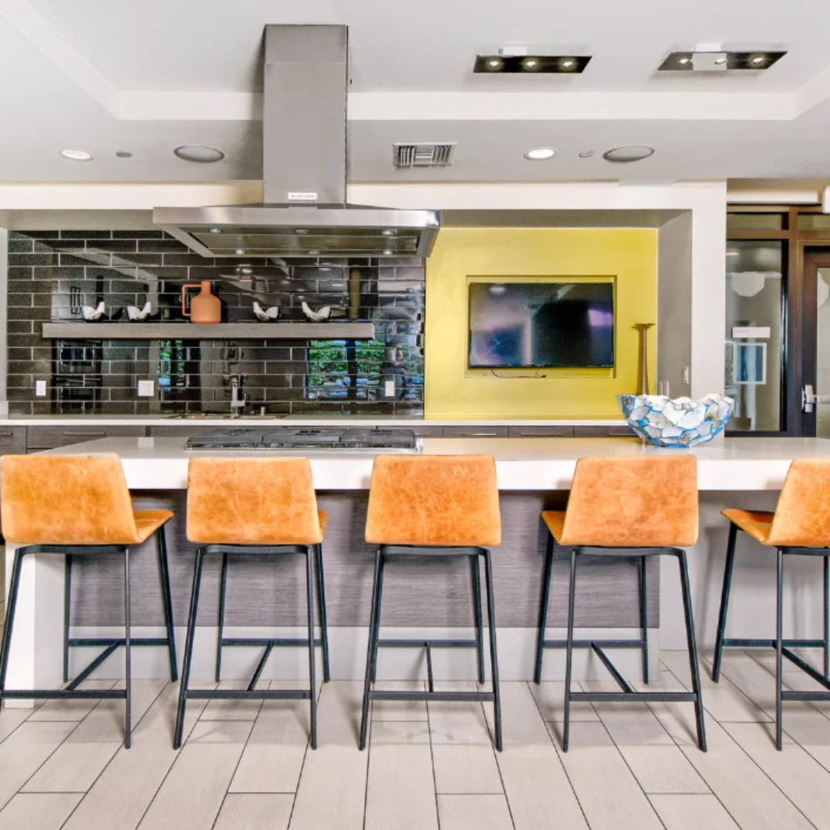 A modern kitchen features a large island with five orange bar stools and stainless steel appliances against a gray and black tiled backsplash.