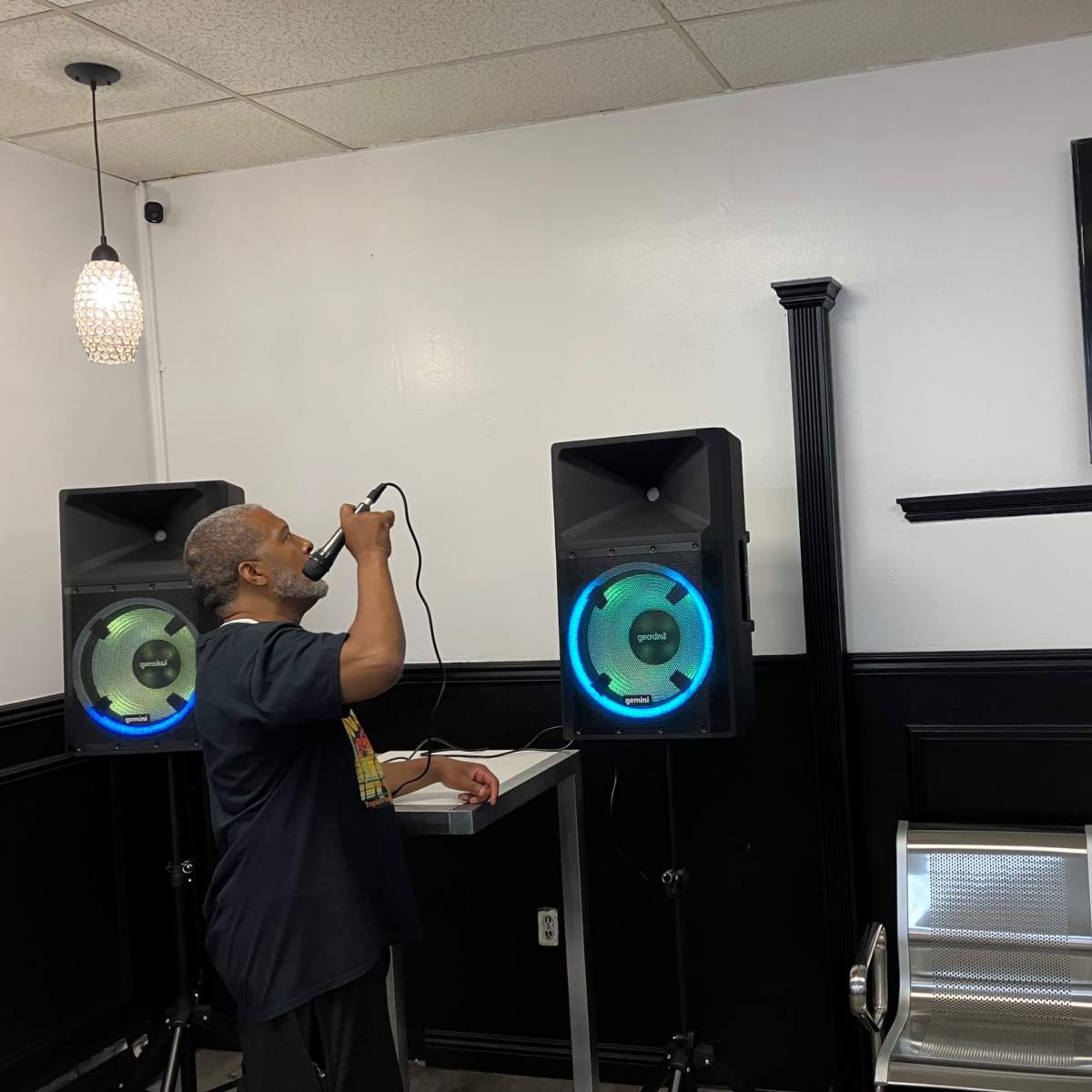 A man stands in front of two speakers, holding a microphone while looking up at a television mounted on the wall.