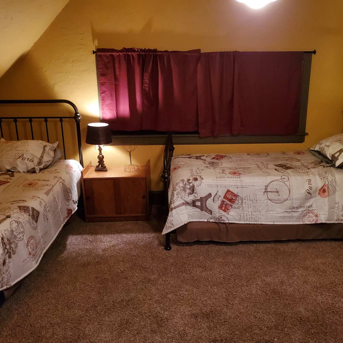 A cozy attic room with two twin beds, a bedside lamp, and a window covered by burgundy curtains.
