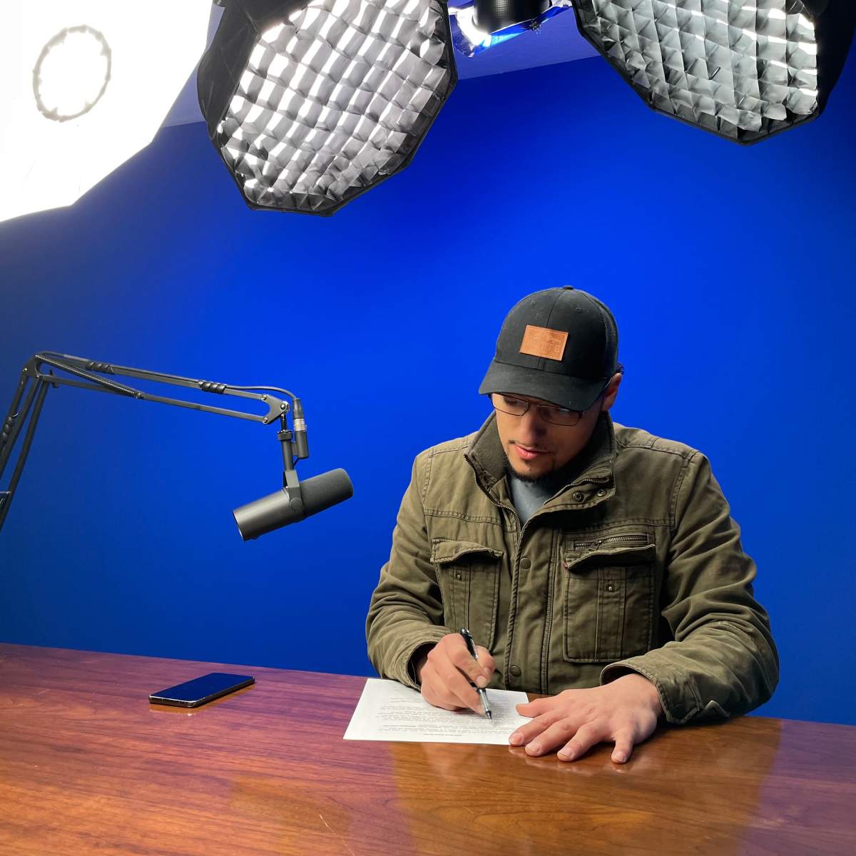 A person wearing a cap writes on paper at a wooden table under bright studio lights, with a blue backdrop and a microphone positioned nearby.