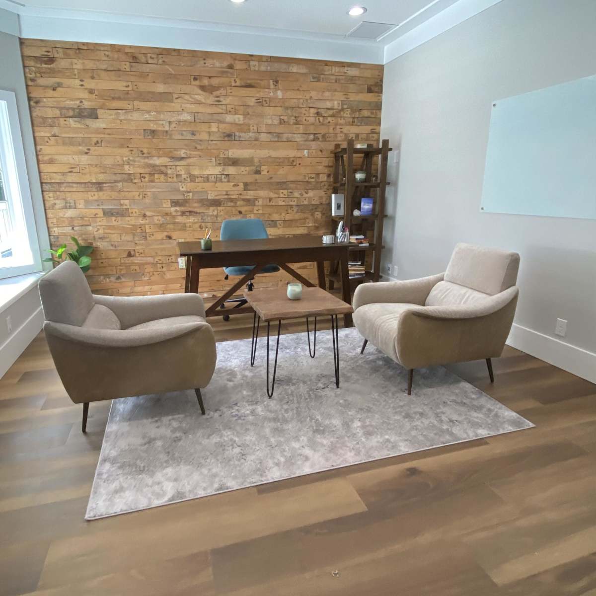 A modern office with a wooden accent wall, a desk in the center, and two armchairs positioned around a small coffee table on a light area rug.