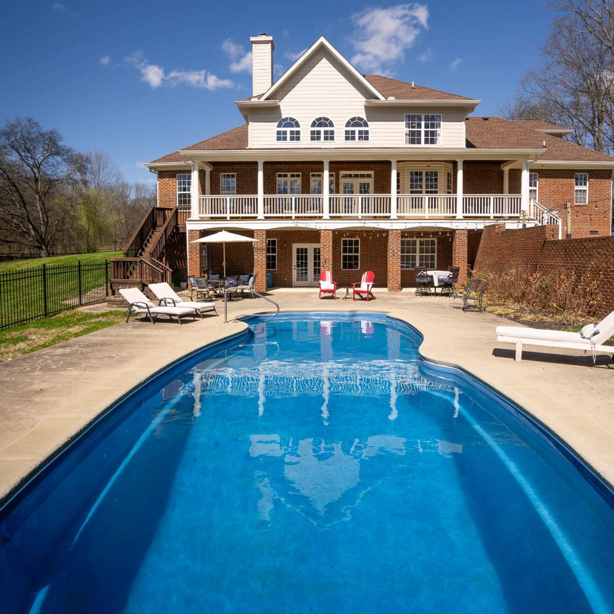 A large brick house with multiple levels overlooks a blue swimming pool surrounded by lounge chairs.