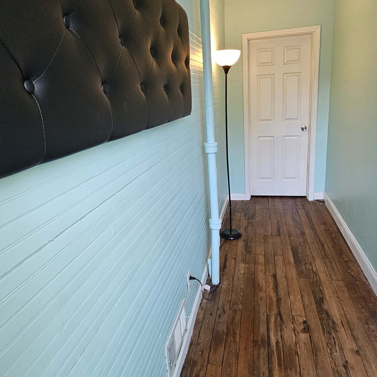 A narrow hallway with light blue walls, a tufted black panel on one side, a standing lamp, and a closed white door at the end.