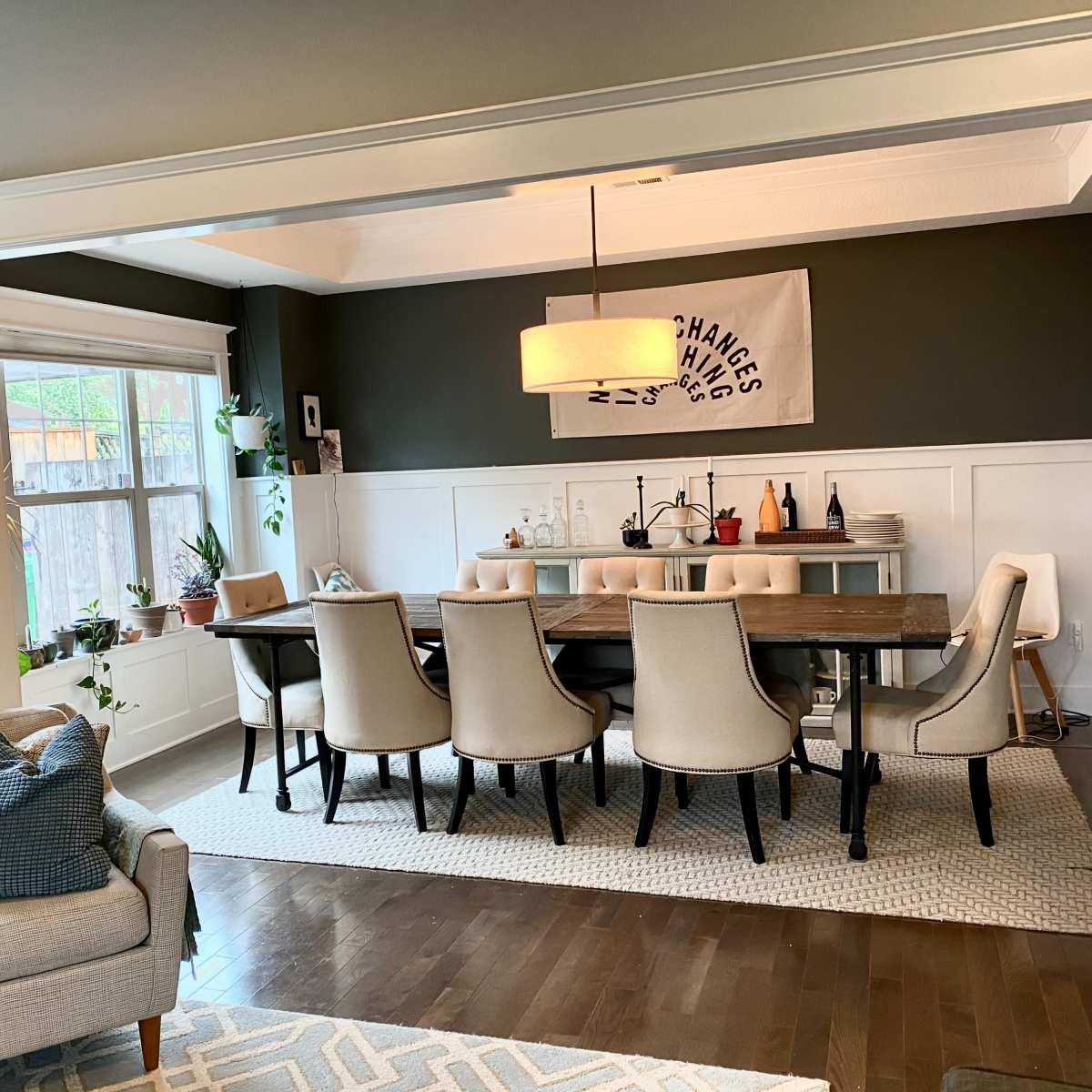 A dining area features a long table surrounded by beige chairs, with a modern light fixture above and decorative items on a side table.