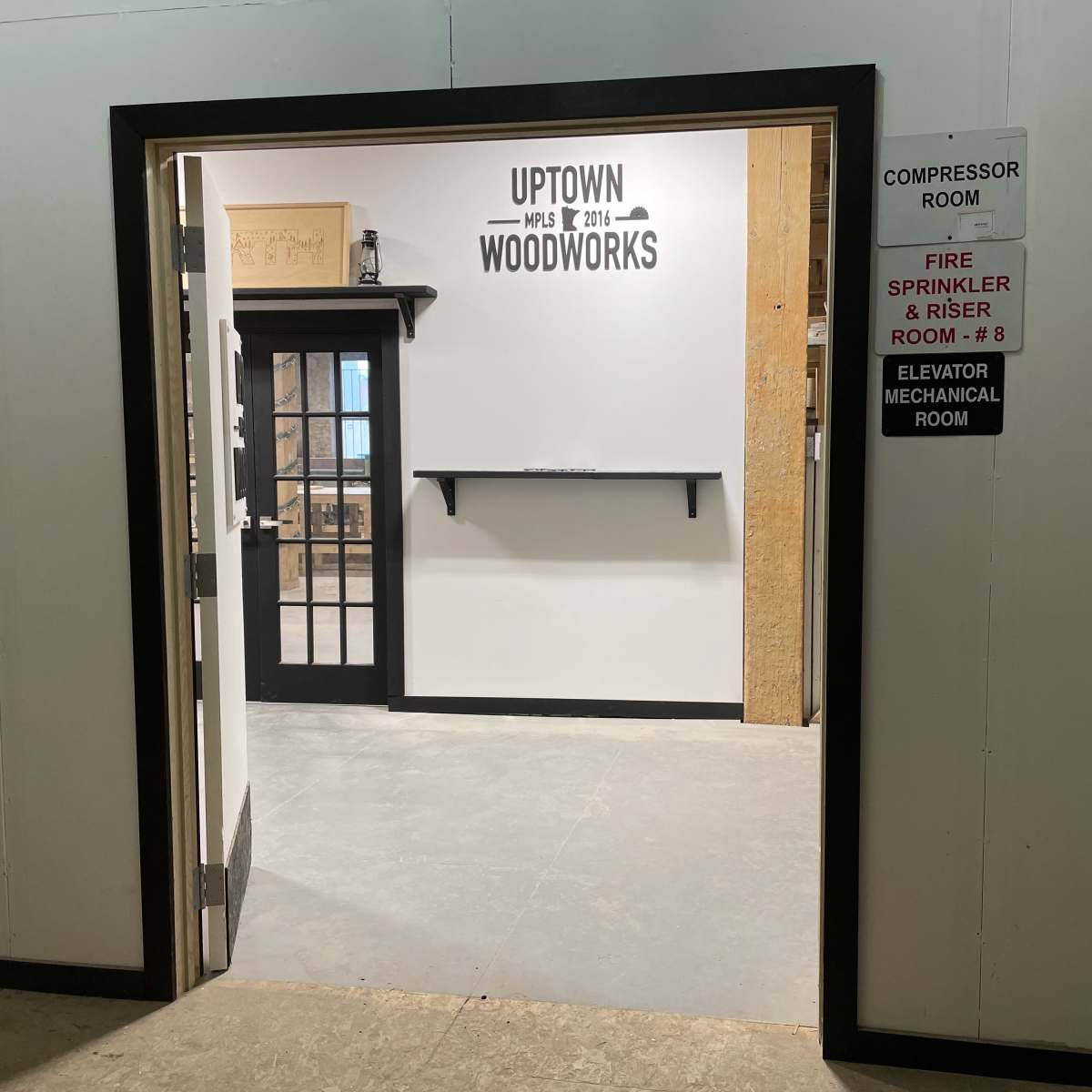 An open doorway leading into a workshop area with a sign reading "Uptown Woodworks" on the wall and a shelf mounted beside the entrance.