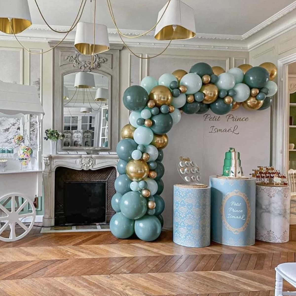A decorated indoor space featuring a balloon arch in shades of teal and gold, a small beverage cart, and a dessert table.