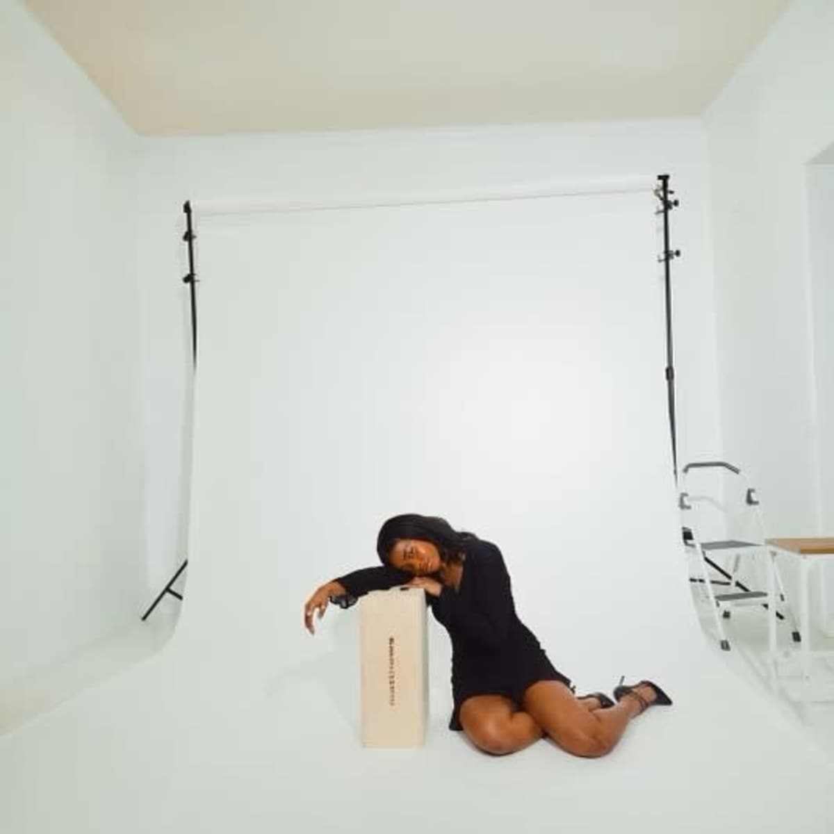 A woman in a black dress is kneeling beside a wooden box in a studio with a white backdrop.