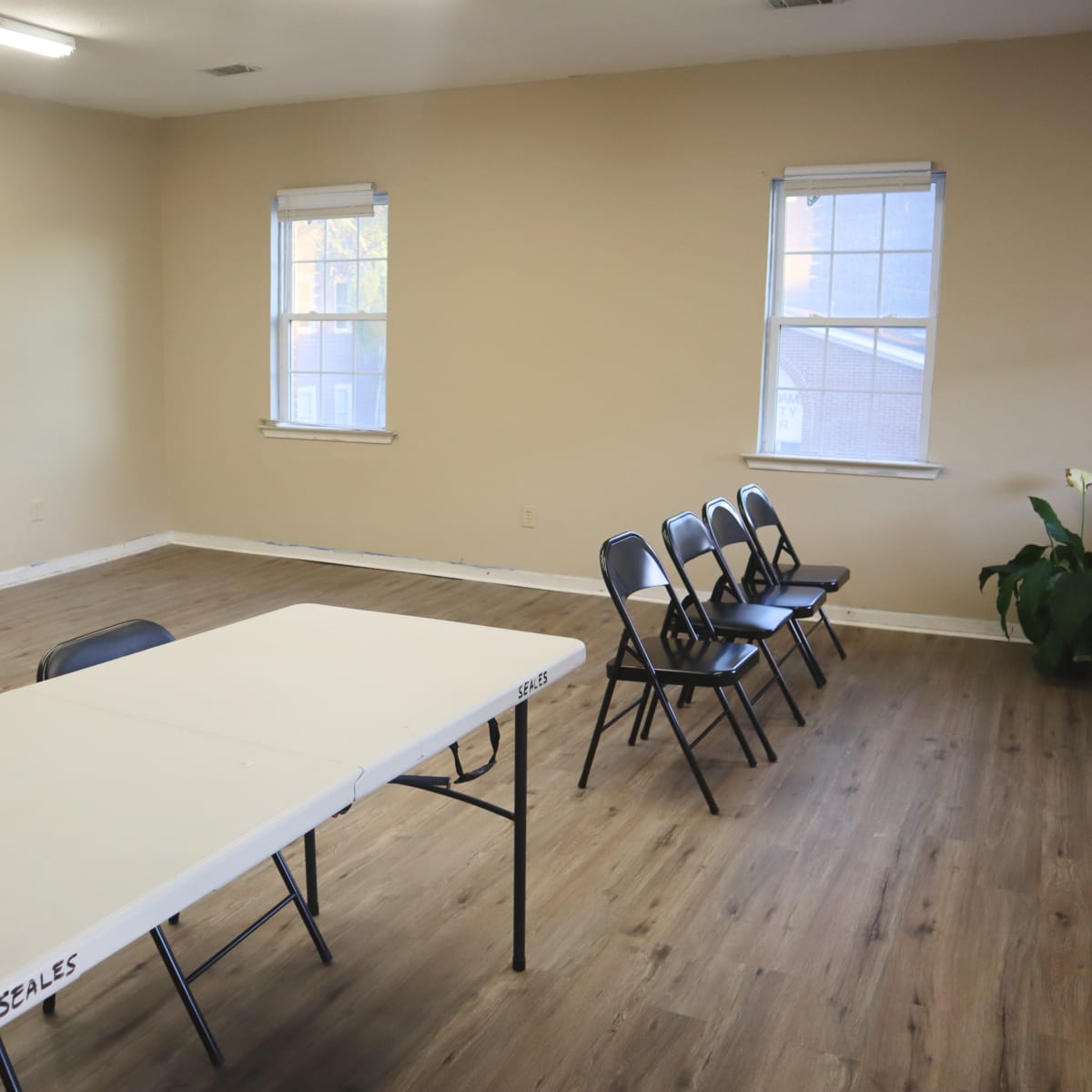 A sparsely furnished room with a long table and several chairs arranged in front of two windows.
